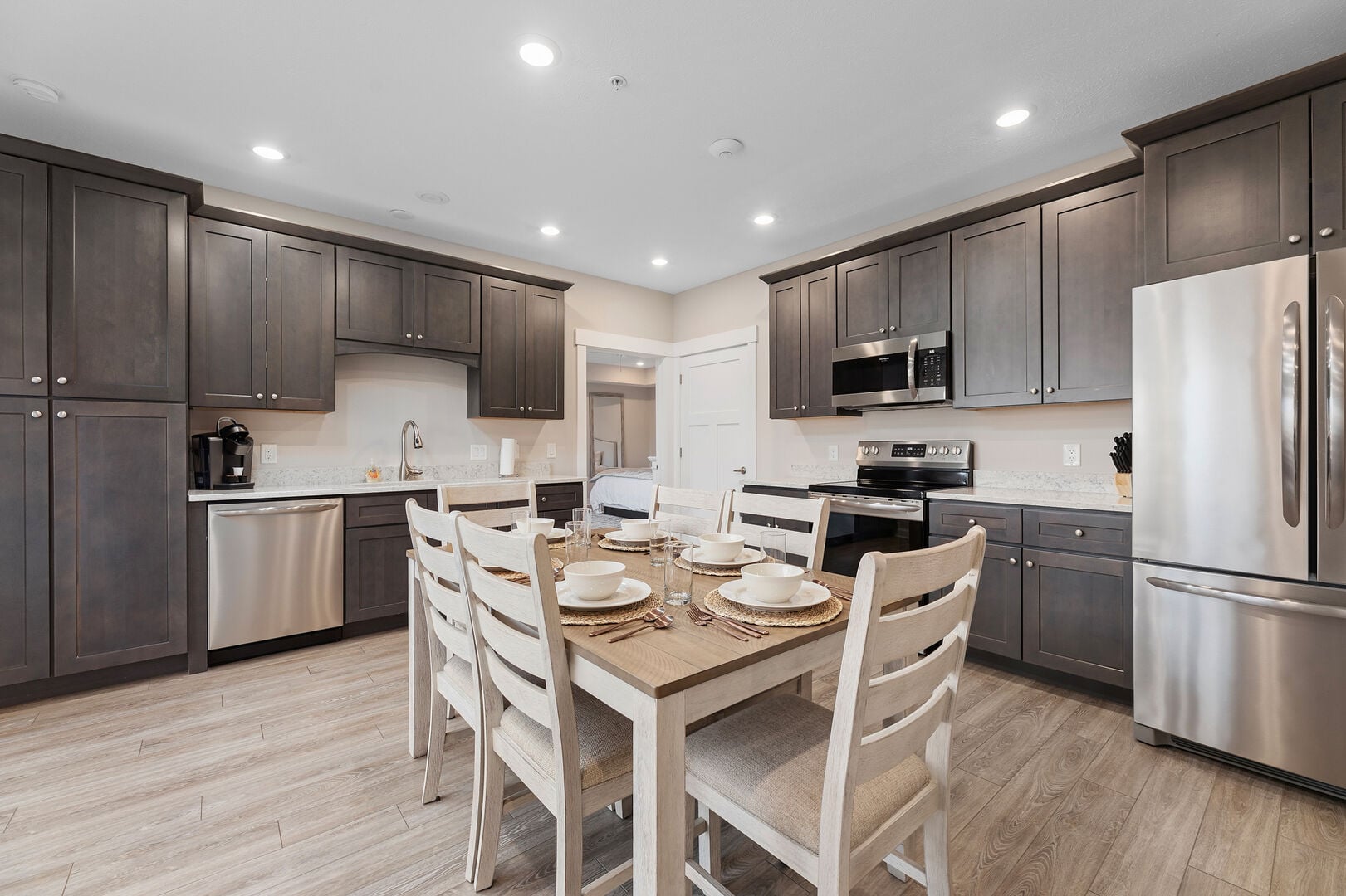 Wooden dining table with white dishes surrounded by dark cabinets and stainless kitchen appliances